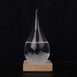 Goccia di vetro trasparente con liquido e bolle, posizionata su base di legno, perfetta come decorazione scientifica o oggetto da collezione.