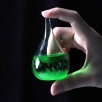 Flacone di laboratorio con liquido fluorescente verde acceso, tenuto tra le dita di una mano, sfondo scuro.