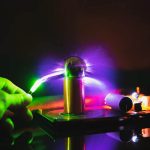 Immagine di una pila di Tesla con scarica elettrica colorata, illuminata da luci viola, verdi e blu, che evidenzia l'effetto di alta tensione e fenomeni elettrici.