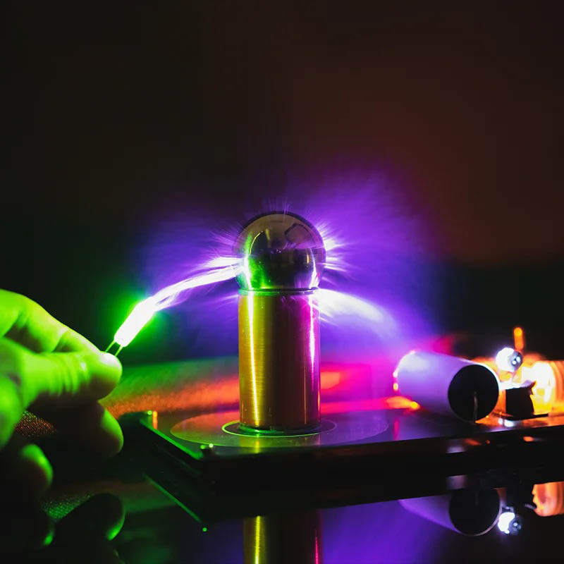 Pila di Tesla con scarica elettrica colorata Immagine di una pila di Tesla con scarica elettrica colorata, illuminata da luci viola, verdi e blu, che evidenzia l'effetto di alta tensione e fenomeni elettrici.