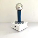 Pila di Van de Graaff con sfera metallica riflettente e base bianca, utilizzata per esperimenti di elettrostatica e decorazioni scientifiche.