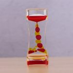 Orologio a clessidra decorativa con sfere rosse e gialle, perfetto come oggetto di arredamento scientifico e decorativo.