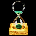 Clessidra decorativa con sabbia verde fluorescente che scorre, posizionata su base di legno naturale, ideale come oggetto scientifico e decorativo.