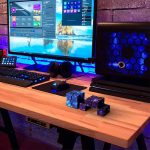 Setup gaming con monitor, tastiera, mouse e cubi di Rubik's Cube luminosi su scrivania in legno, illuminato da luci blu e rosa, ambiente moderno e tecnologico.