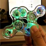 Fidget spinner con luci LED verdi e blu, oggetto scientifico decorativo, maneggiato da una mano, sfondo di uno schermo e ambiente interno.