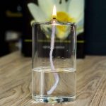 Fiammella di candela accesa in un bicchiere d'acqua, con un fiore sfocato sullo sfondo, creando un'immagine suggestiva e decorativa.