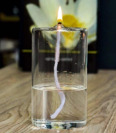 Fiammella di candela accesa in un bicchiere d'acqua, con un fiore sfocato sullo sfondo, creando un'immagine suggestiva e decorativa.