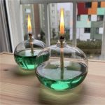 Due oggetti di vetro a forma di sfera con liquido verde e fiamme accese, posizionati su un tavolo di legno vicino a una finestra con vista urbana.