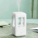 Vaporizzatore portatile bianco con vapore in uscita, ideale per aromaterapia e benessere, posizionato su un tavolo con sfondo verde e tende.