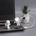 Decorazione scientifica originale con due astronauti in miniatura collegati a una lampada LED, posizionati su un laptop, con pianta succulenta in vaso trasparente sullo sfondo.