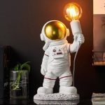 Statua di astronauta con casco dorato e lampada a forma di lampadina accesa, decorazione scientifica originale per ambienti moderni e tematici.