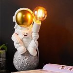 Decorazione di un astronauta con casco dorato che tiene una lampada a forma di lampadina accesa, posizionato su una superficie con decorazioni spaziali.