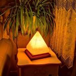 Lampada decorativa a forma di piramide illuminata, posizionata su un tavolino vicino a una pianta e a una tenda ricamata, in un ambiente caldo e accogliente.
