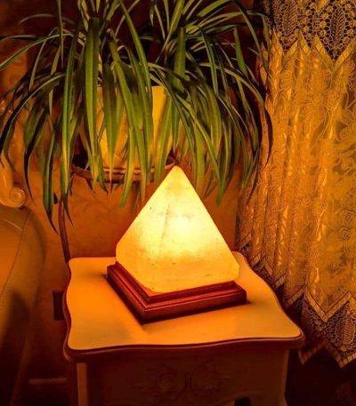 Lampada decorativa a forma di piramide illuminata, posizionata su un tavolino vicino a una pianta e a una tenda ricamata, in un ambiente caldo e accogliente.