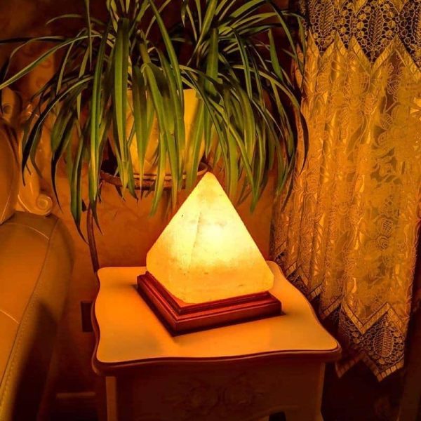 Lampada decorativa a forma di piramide illuminata, posizionata su un tavolino vicino a una pianta e a una tenda ricamata, in un ambiente caldo e accogliente.