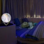 Sfera luminosa decorativa con motivi scientifici, posizionata su un supporto, in un ambiente con luci soffuse e tende, creando un'atmosfera accogliente e stimolante.