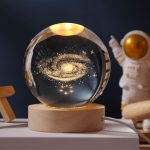 Vaso di cristallo con rappresentazione di una galassia luminosa e dettagli stellari, posizionato su una base di legno, con sfondo scuro.