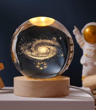 Vaso di cristallo con rappresentazione di una galassia luminosa e dettagli stellari, posizionato su una base di legno, con sfondo scuro.