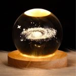 Sfera di cristallo con rappresentazione di una galassia, decorata con stelle e nebulose, posizionata su un supporto di legno.