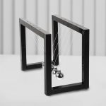 Péndolo di Newton con cornice nera e sfondo bianco, mostra le sfere metalliche che oscillano, illustrando i principi della fisica e della conservazione dell'energia.
