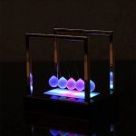 Pendo di Newton illuminato con luci LED colorate, oggetto decorativo scientifico con sfere sospese in un supporto nero, ideale per decorazioni moderne e educative.