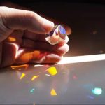 Mano che tiene una pietra di cristallo trasparente che proietta riflessi di luce colorati su una superficie bianca, creando effetti di luce e colore.