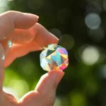 Mani che tengono una palla di cristallo iridescente con riflessi colorati, sfondo naturale sfocato.