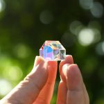Oggetto scientifico in vetro colorato, trasparente e riflettente, tenuto tra le dita con sfondo naturale sfocato.