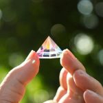 Mani che tengono una piccola piramide di cristallo trasparente, riflessi di luce e sfondo naturale sfocato.