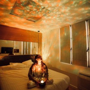 Donna seduta sul letto in una stanza con decorazioni a tema astronomico, proiettando stelle e pianeti sul soffitto e le pareti, creando un'atmosfera magica e rilassante.
