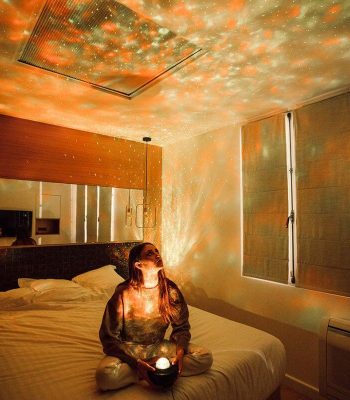 Donna seduta sul letto in una stanza con decorazioni a tema astronomico, proiettando stelle e pianeti sul soffitto e le pareti, creando un'atmosfera magica e rilassante.