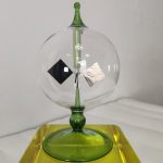 Vaso di cristallo trasparente con oggetti scientifici decorativi, posizionato su base verde e sfondo neutro. Perfetto come decorazione per appassionati di scienza e design.