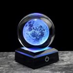 Modello di luna in cristallo illuminata con luce blu, posizionata su base nera con pulsante di accensione, perfetta come decorazione scientifica o oggetto da collezione.