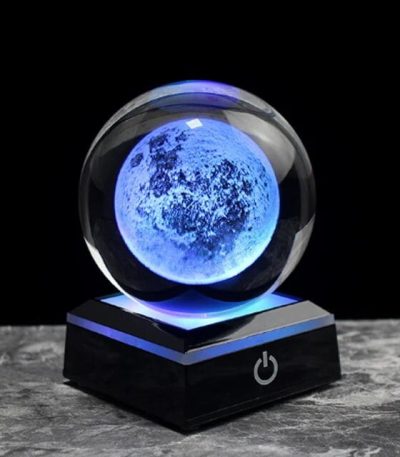 Modello di luna in cristallo illuminata con luce blu, posizionata su base nera con pulsante di accensione, perfetta come decorazione scientifica o oggetto da collezione.