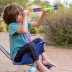 Bambina che guarda attraverso un telescopio colorato all'aperto, seduta su una sedia blu, in un ambiente naturale con piante e cielo azzurro.