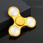 Fidget spinner dorato con tre bracci e centro rotante, oggetto di gioco e relax, posizionato su superficie nera con scatola nera sullo sfondo.