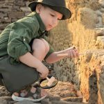 Pezzo di testo 2: Bambino curioso che osserva attentamente i reperti archeologici con una lente d'ingrandimento, indossando abbigliamento da esploratore e cappello, in un sito storico all'aperto.