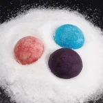 Palline di coloranti in polvere di vari colori (rosa, blu, viola) disposte su una superficie di sale bianco, ideali per decorazioni scientifiche e attività educative.