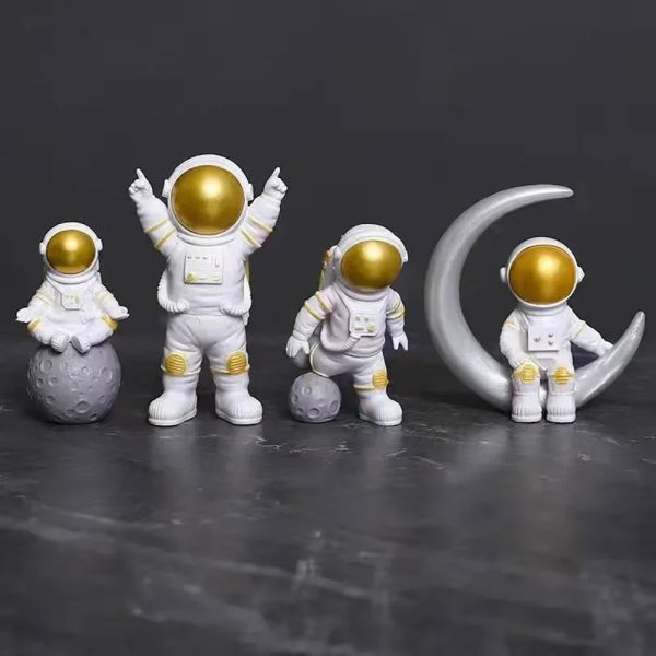 Astronauti in miniatura con caschi dorati e tute bianche, posizionati su una superficie scura, con uno seduto sulla luna e altri in piedi, creando un'atmosfera spaziale e futuristica.
