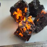 Immagine di un meteorite con superficie irregolare e lucente, che mostra dettagli di texture e colori scuri con zone luminose arancioni, posizionato su una superficie chiara con una scala di misura.
