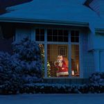 Babbo Natale vestito con abiti tradizionali natalizi, visibile attraverso la finestra di una casa, con decorazioni festive e un albero di Natale illuminato all’interno.