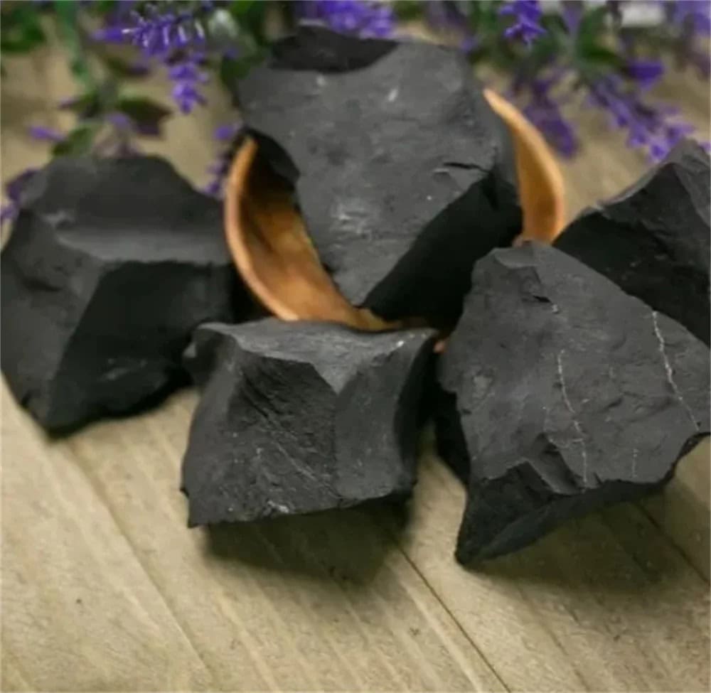 Pietre di carbonio naturale nere, perfette per decorazioni scientifiche e progetti educativi, disposte su un tavolo di legno con fiori viola sullo sfondo.