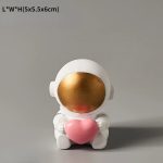 Statua decorativa di un astronauta con viso dorato e grande cuore rosa, ideale come decorazione per appassionati di scienza e spazio. Perfetto come regalo originale o elemento di arredo tematico.