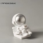 Statua di un astronauta in stile scultoreo, con casco argentato e tuta bianca, seduto su una base. Perfetto come decorazione scientifica o da collezione.