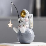 Figurina di astronauta in tuta spaziale seduto su una luna, che tiene una canna da pesca con una stella appesa, decorazione scientifica originale e fantasiosa.