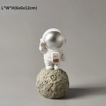 Vaso decorativo a forma di astronauta che saluta, posizionato su una superficie che ricorda la luna, perfetto come oggetto decorativo scientifico o da collezione.