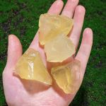 Quattro grandi cristalli di gesso naturale, traslucidi e giallastri, tenuti in mano contro uno sfondo di erba verde.