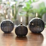 Tre palline di vetro nere con candele accese, posizionate su un tavolo di legno, con sfondo di piante verdi e luce naturale.