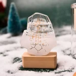 Orologio artistico in vetro trasparente con disegni scientifici, posizionato su base di legno, ambientato in un paesaggio invernale con neve e alberi sfocati sullo sfondo.
