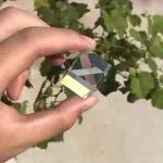 Cristallo decorativo trasparente con riflessi arcobaleno, tenuto tra le dita di una mano, con sfondo di foglie verdi.